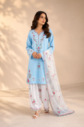 A woman models a light blue and white traditional South Asian outfit with floral embroidery and a matching sheer dupatta, set against a warm beige background with soft leaf shadows.