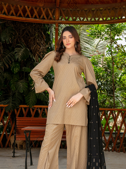 A woman modeling a beige three-piece salwar kameez suit with matching wide-leg trousers and a black dupatta with small white motifs, posed under a wooden pavilion with lush green foliage in the background.