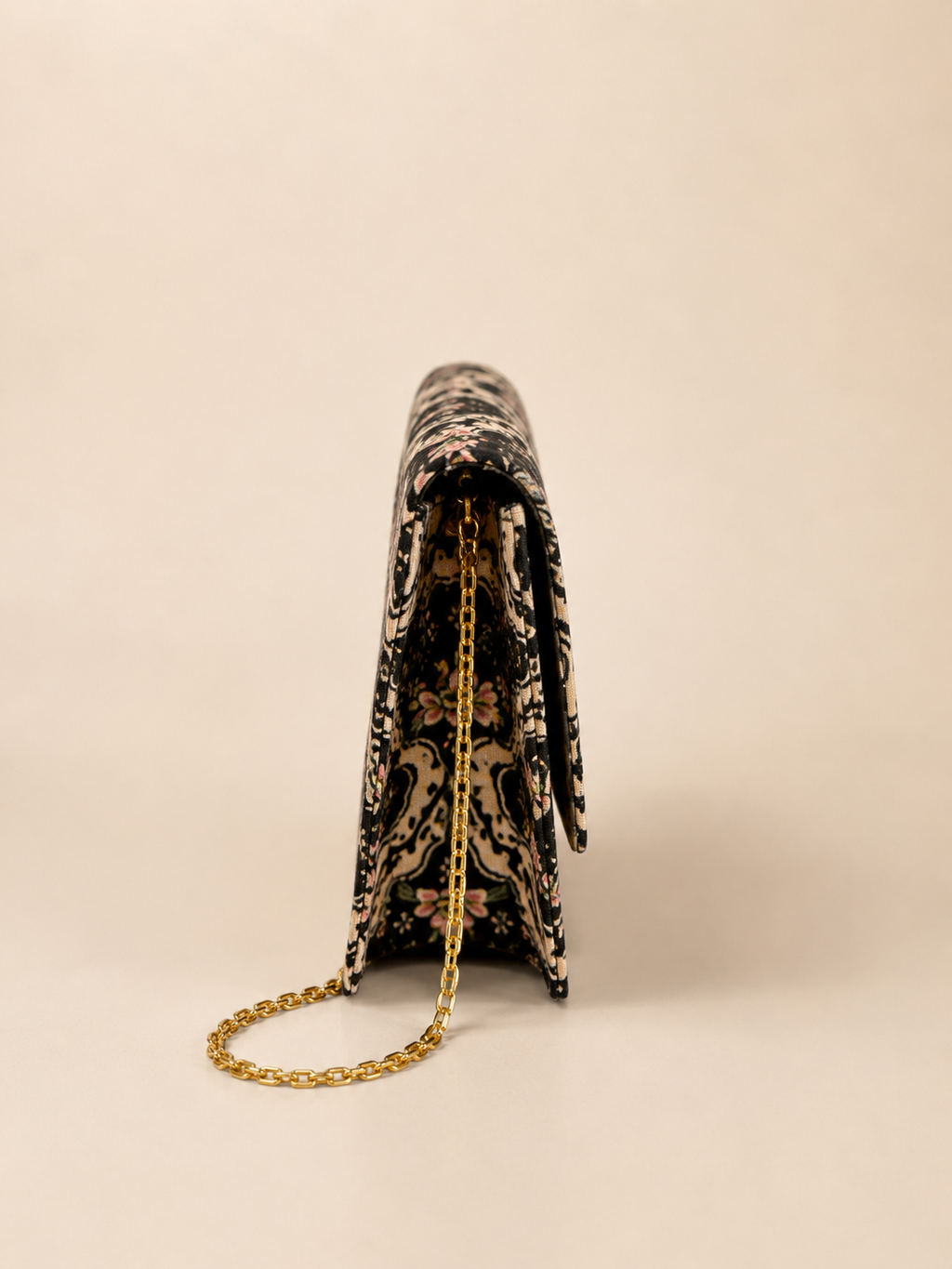 A black and beige paisley patterned clutch bag with a gold chain strap, shown from a side profile view against a solid beige background.