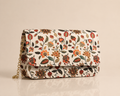 A rectangular clutch bag with a cream background and an all-over floral pattern in orange, red, green, and brown. It features a gold chain strap on the left side, set against a solid beige background with a soft reflection below.