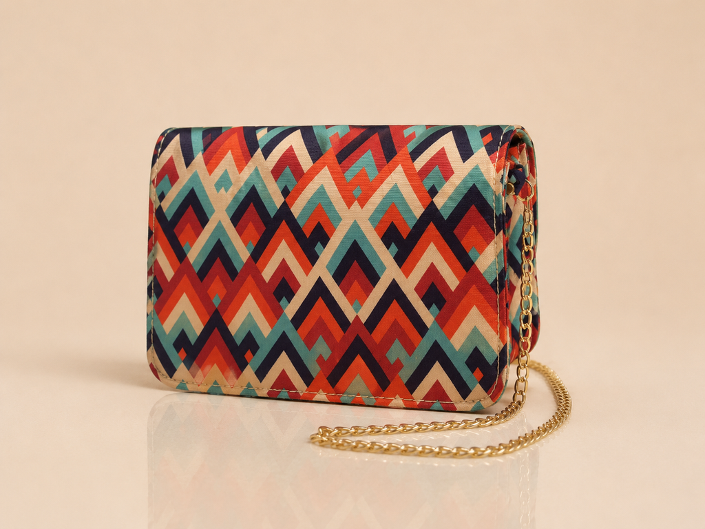 A small rectangular crossbody bag with a colorful geometric chevron pattern in red, orange, teal, and cream, featuring a gold chain strap, set against a warm beige background.