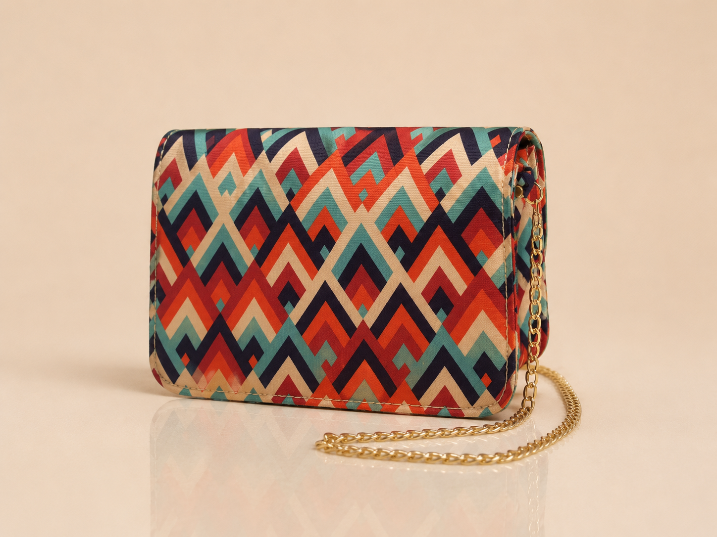 A small rectangular crossbody bag with a colorful geometric chevron pattern in red, orange, teal, and cream, featuring a gold chain strap, set against a warm beige background.